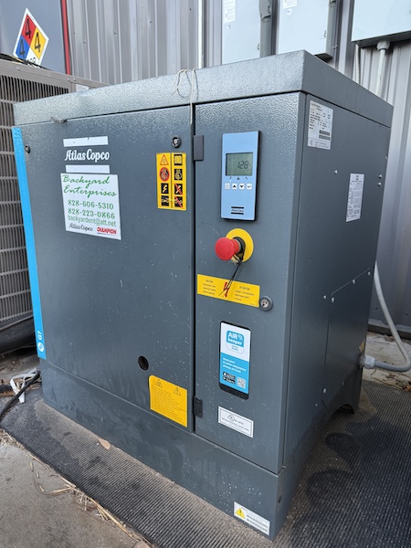 An Atlas Copco compressor installed outdoors, its controller display showing the running pressure and a service company sticker on the door.