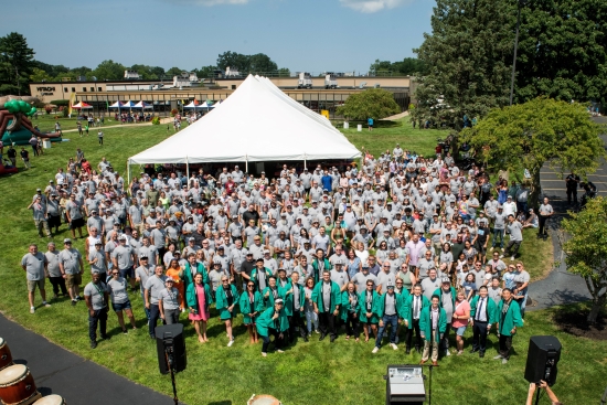 An aerial group photograph of several hundred employees assembled on a lawn in front of a marquee.
