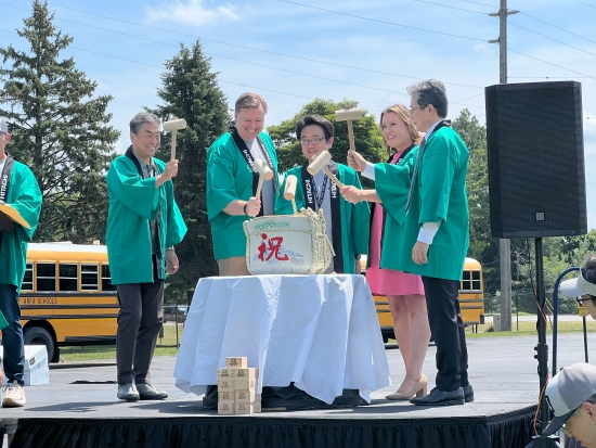 A sake barrel breaking ceremony on an outdoor stage, participants in green happi coats raising mallets over a decorated barrel.