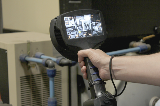 A hand holding an acoustic imaging camera in front of compressed air pipework, the leak located on its screen.
