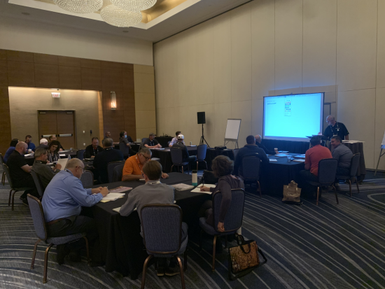 A training session in progress, delegates seated at round tables facing an instructor and a projection screen.