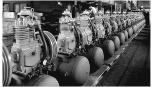 Historic black and white photograph of reciprocating compressors lined up along an assembly line.