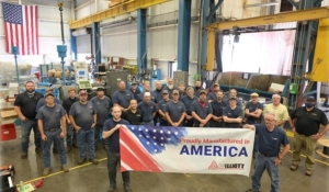 Factory staff assembled beneath a Proudly Manufactured in America banner in an assembly bay.