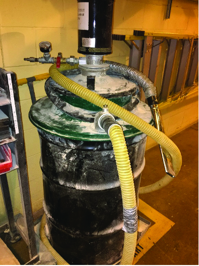 A drum with a filter head and flexible hoses connected, standing against a plant wall.