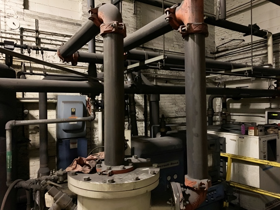 Foundry compressed air piping before rework, with large vertical risers rising from a flanged base into an overhead header in a basement plant room.