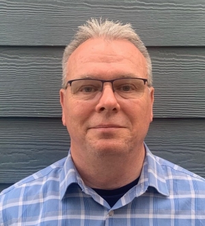 A man in a checked shirt photographed against a clapboard wall.