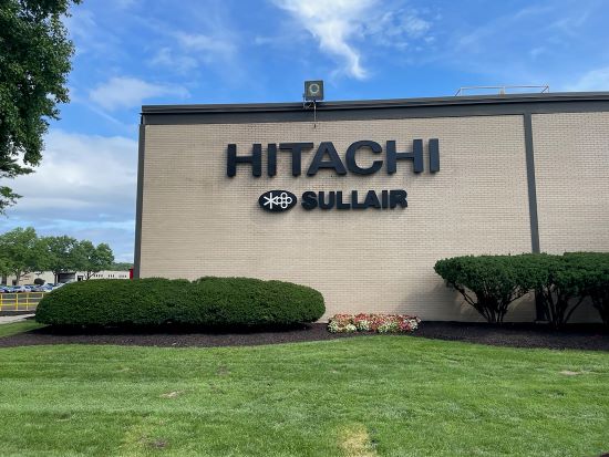 A building exterior with signage reading Hitachi Sullair, above trimmed shrubs and a lawn.