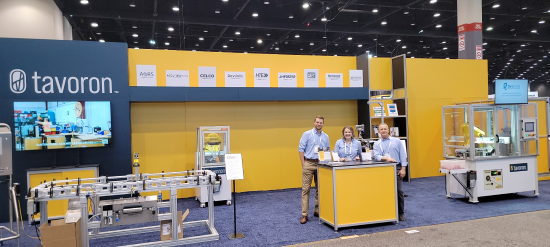 A tavoron exhibition stand with three people at the counter, packaging machinery displayed to either side and partner logos along the back wall.