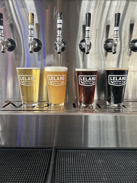 Four glasses of beer of different colours lined up on a bar in front of the taps, each branded Leland Brewing Co.