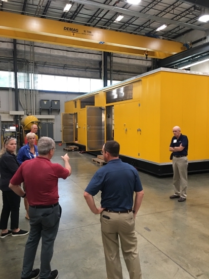 A factory tour group standing beside large yellow compressor enclosures under an overhead crane.