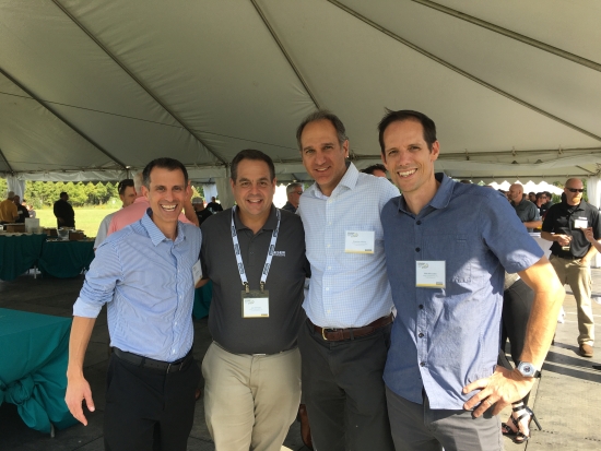 Four men photographed together under a marquee at an outdoor event.