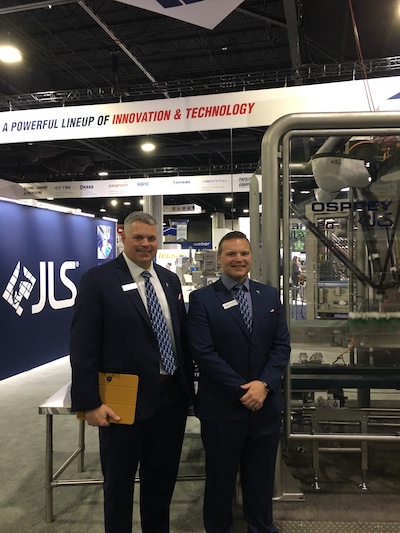 Two men in suits standing in front of packaging machinery on a trade show stand.