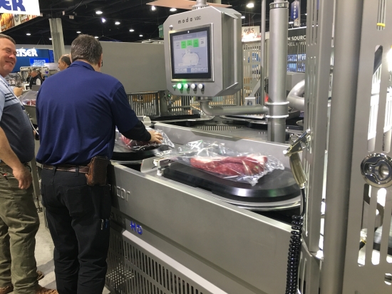 A man operating a vacuum packaging machine at a trade show, a tray of meat sealed under film on the belt.