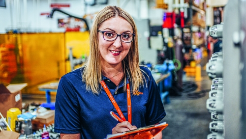 A woman in a company polo shirt holding a clipboard in a parts workshop.