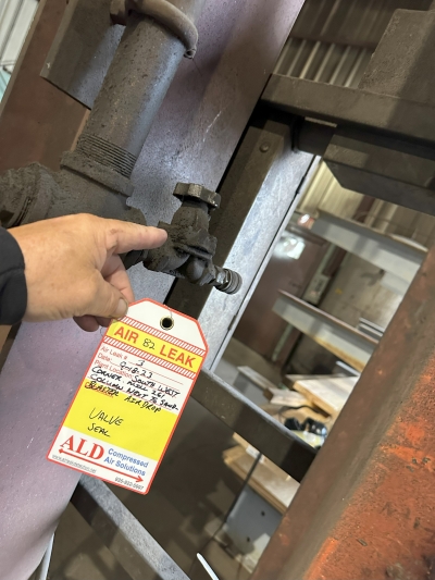 A hand pointing to a leaking valve seal, with a compressed air leak tag attached recording the location, size and estimated cost.
