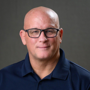 A man in a dark polo shirt photographed against a grey background.