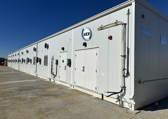 A long white modular equipment building on a concrete pad under a clear sky.