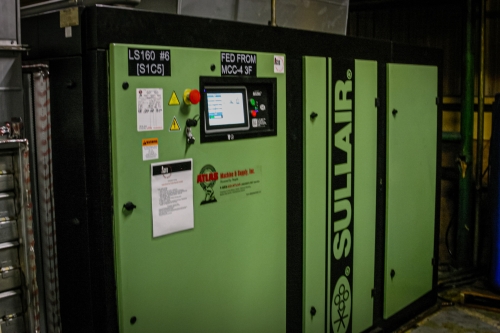 A green Sullair compressor in a plant room, its cabinet labelled with the unit number and the motor control centre it is fed from.
