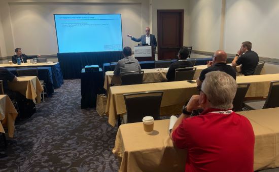 A conference session in a hotel meeting room, a presenter at the lectern beside a projected slide.