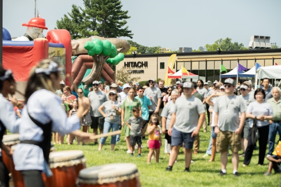 A company family day on a lawn outside a Hitachi building, taiko drummers in the foreground and a crowd watching.