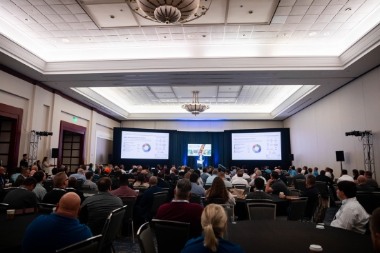 A conference session in a hotel ballroom, delegates seated at round tables facing two projection screens.