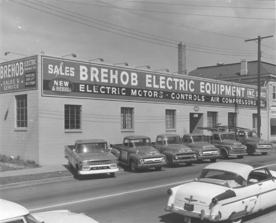 Historic photograph of the Brehob Electric Equipment building, its signage advertising electric motors, controls and air compressors, with a row of pickup trucks parked outside.